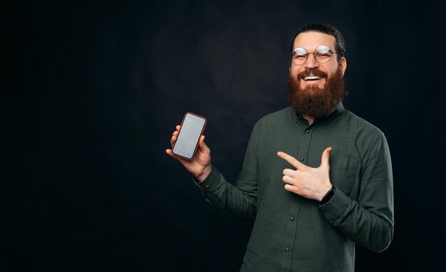 Un homme barbu paisible portant des lunettes pointe vers le téléphone qu'il tient dans un studio sur fond noir