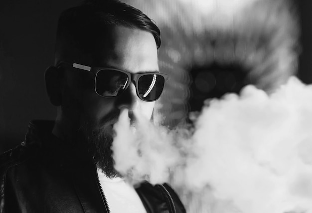 Photo homme barbu pensif vêtu d'une veste en cuir lunettes de soleil vaping un nuage de vapeur