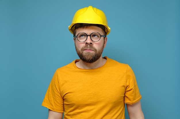 Homme barbu perplexe dans un casque de construction et des lunettes rondes lève les yeux pensivement