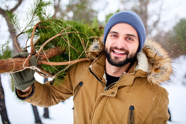 Photo homme barbu portant un arbre de noël fraîchement coupé dans la forêt jeune bûcheron porte sapin sur