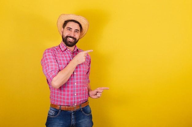 Homme barbu portant des vêtements typiques pour la fête Junina montrant avec les doigts sur le côté la publicité négative et l'espace de texte pour la fête d'Arraia