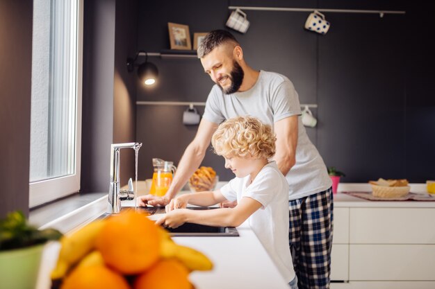 Homme barbu positif aidant son fils à laver la vaisselle