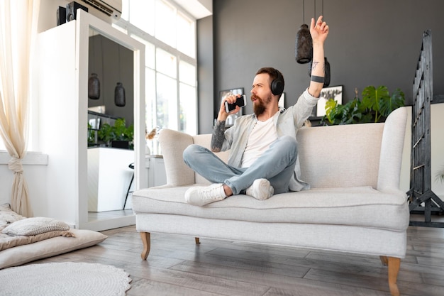 Homme barbu positif écoutant de la musique avec des écouteurs tout en étant assis sur un canapé à la maison