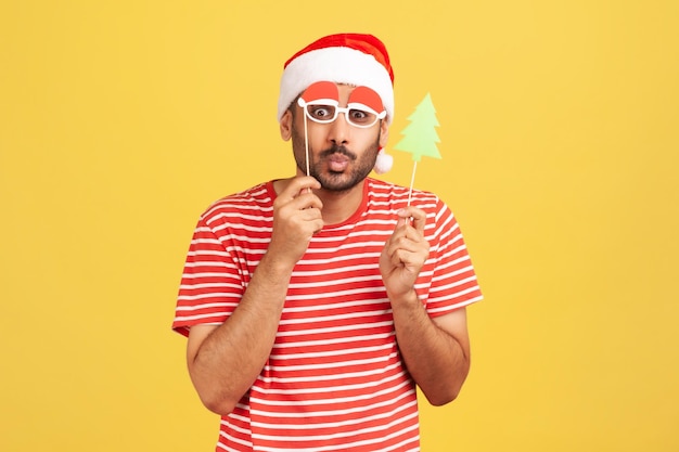 Homme barbu positif en t-shirt rouge et en chapeau de père Noël tromper les visages s'amusant à essayer des masques de fête de Noël sur bâton Prise de vue en studio intérieure isolée sur fond jaune