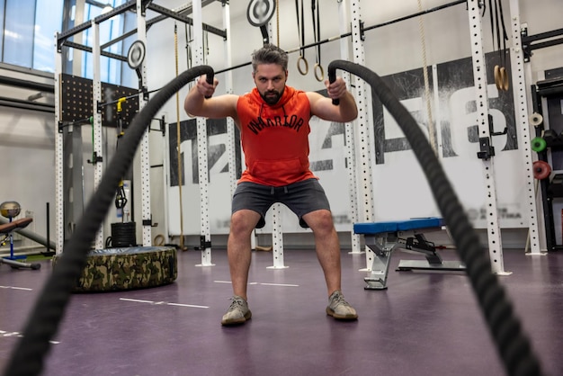 Photo un homme barbu puissant et fort s'entraîne avec des cordes de combat au gymnase pour développer la force et l'endurance.