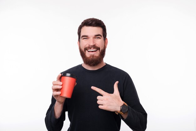 Homme barbu en pull noir tient une tasse orange et la pointe sur un mur blanc.