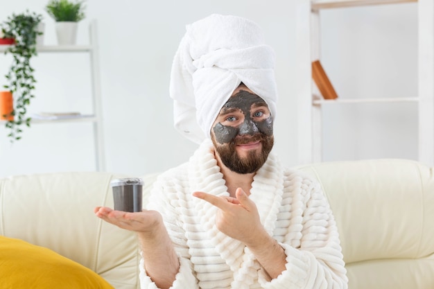 Homme barbu s'amusant avec un masque cosmétique sur son visage fait d'humour de soins de la peau pour hommes d'argile noire et