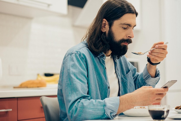 Homme barbu séduisant assis à la table et mangeant des cornflakes tout en regardant l'écran d'un smartphone moderne