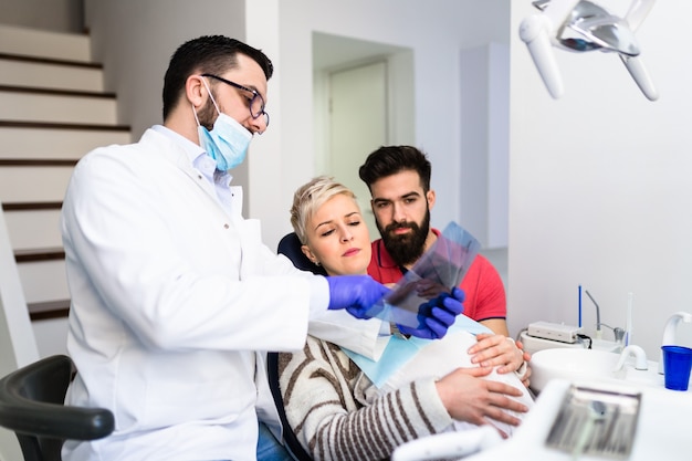 Homme barbu séduisant avec sa femme enceinte recevant un traitement médical dans un cabinet dentaire.