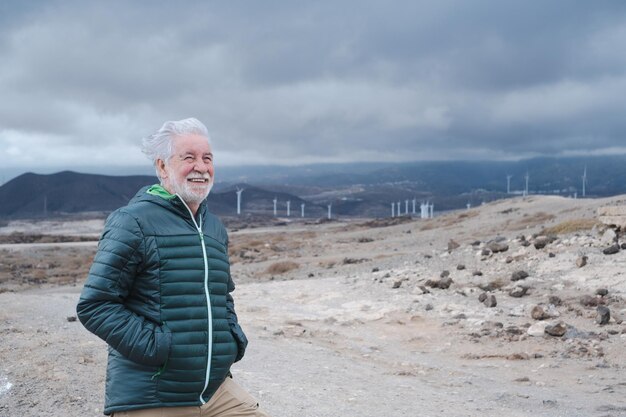 Homme barbu senior souriant en plein air par une journée nuageuse venteuse. Ferme éolienne sur fond