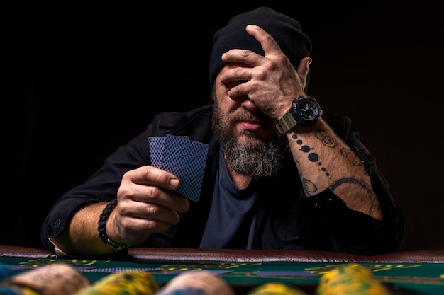 Homme barbu sérieux assis à une table de poker et tenant des cartes isolées sur fond noir