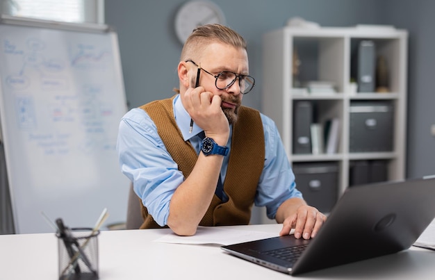 Homme barbu sérieux dans des lunettes à l'aide d'un ordinateur portable sans fil assis à table