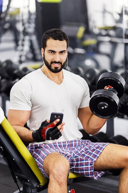 Un homme barbu sexy et fort porte un t-shirt blanc et des écouteurs tient le téléphone et l'haltère dans la salle de sport