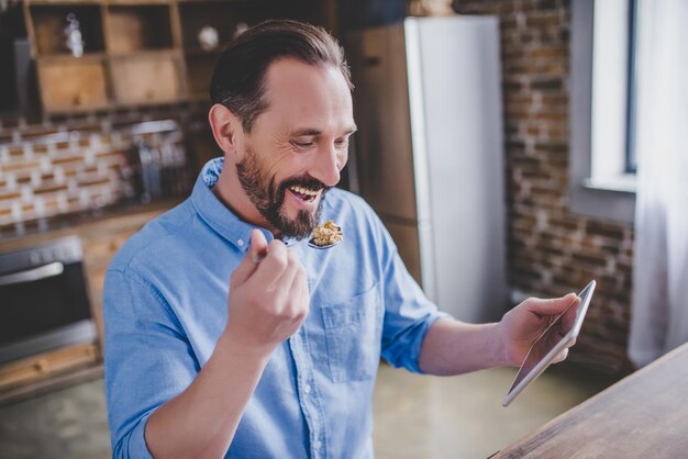 Homme barbu souriant mangeant des céréales pour le petit-déjeuner et utilisant une tablette numérique dans la cuisine