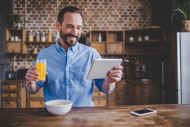 Homme barbu souriant tenant un verre de jus d'orange pour le petit-déjeuner et utilisant une tablette numérique dans la cuisine