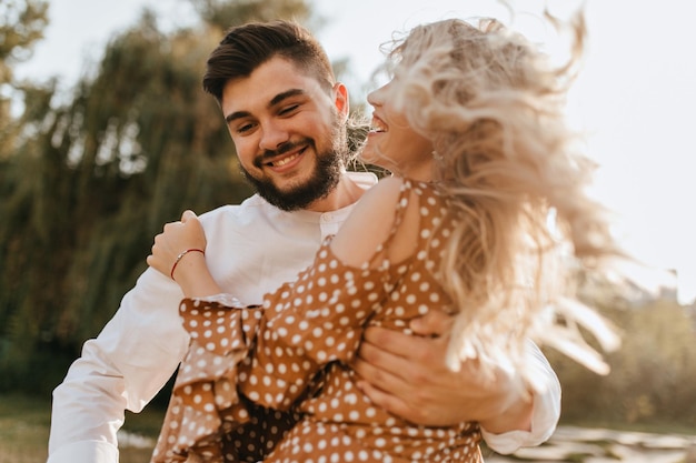 Un homme barbu avec un sourire embrasse une fille bien-aimée positive en chemisier à pois marron