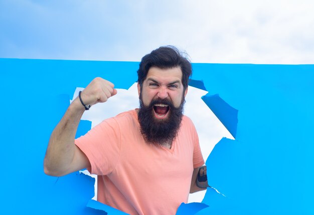 Homme barbu de succès furtivement du trou de papier déchiré homme barbu heureux à travers le trou dans le papier bleu gagner