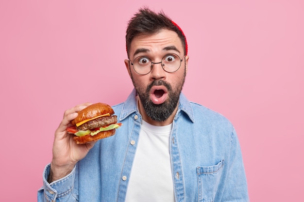 Un homme barbu surpris mange des aliments riches en calories un délicieux hamburger apprécie le goût d'un hamburger frais vêtu de vêtements décontractés