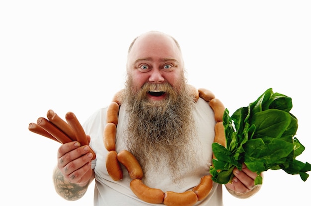 Un homme barbu surweigt en riant portant un bruch de saucisses autour du cou montrant des saucisses et des verts à la caméra. Portrait isolé sur fond blanc.