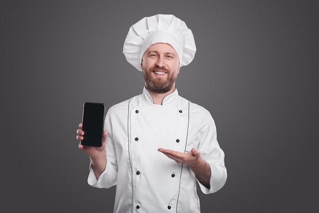 Homme barbu sympathique en uniforme de chef blanc souriant et regardant la caméra tout en faisant de la publicité pour une application mobile sur un smartphone, avec un écran noir vide sur fond gris