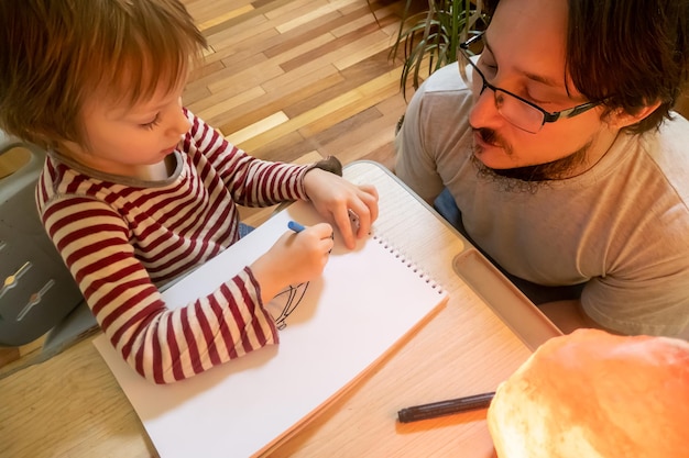 Homme barbu en t-shirt gris et garçon mignon dessinant des images avec des crayons de cire colorés tout en passant du temps ensemble à la maison