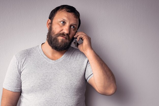 Homme barbu en t-shirt gris tenant un téléphone portable en se tenant debout contre un mur gris