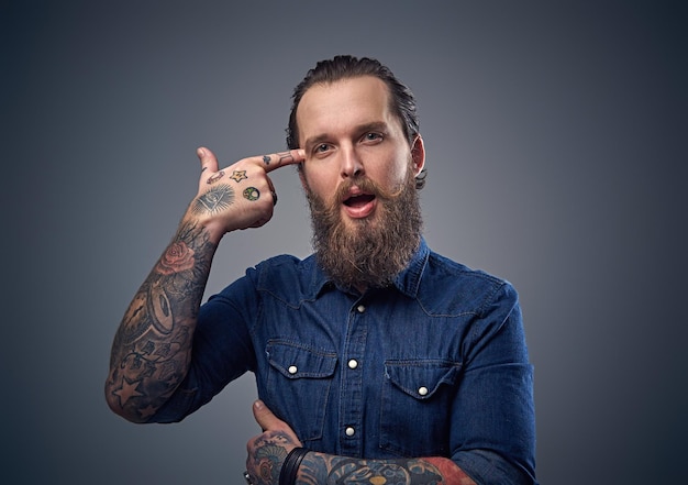 Homme barbu avec des tatouages sur les bras, vêtu d'une chemise en jean sur fond gris.