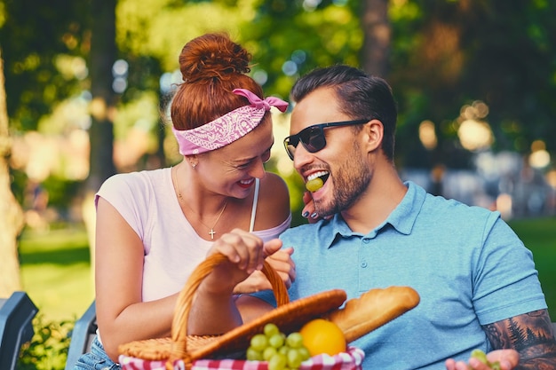 Un homme barbu tatoué et une femme rousse font un pique-nique sur un banc dans un parc.