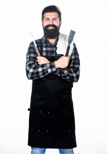 Homme barbu tenant des outils de préhension du gril Hipster en tablier avec des ustensiles en métal pour le gril du barbecue Préparer des aliments sur le gril