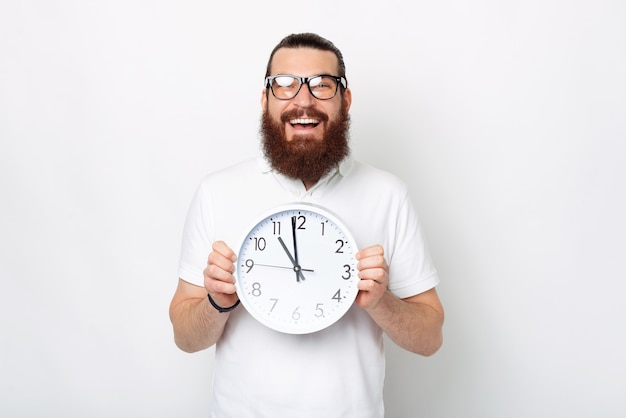 L'homme barbu tient une horloge murale tout en souriant à la caméra.