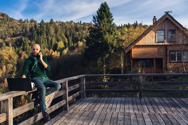 Homme barbu travaille sur un ordinateur portable en parlant au téléphone Hipster indépendant en vacances dans les montagnes