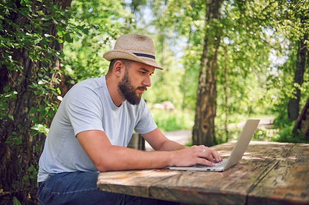 Homme barbu travaille et souriant avec un ordinateur portable dans le parc sous l'arbre. Un pigiste heureux est assis et utilise une application ou un site Web sur l'herbe. Travailler à distance en mode d'auto-isolation