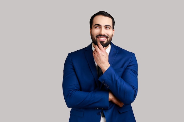 Homme barbu avec un visage paisible réfléchissant à des plans pour l'avenir tenant son menton homme pensif rêvant d'un bon travail portant un costume de style officiel tourné en studio intérieur isolé sur fond gris