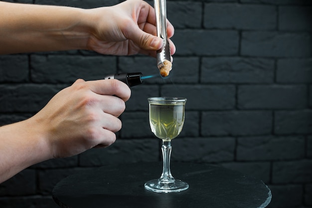 Homme de barman prépare un cocktail alcoolisé chaud dans un bar sur une table en bois noire près du mur de briques à l'aide d'un brûleur professionnel. Délicieux cocktail original.