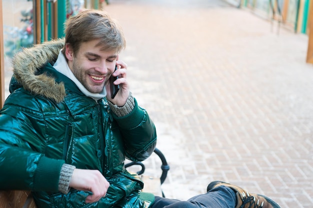 Homme beau tenir smartphone. L'homme non rasé porte une veste chaude et tient un smartphone sur fond urbain enneigé. Guy s'asseoir sur un banc interagit avec un smartphone. Notion de communication. Hipster utilise un smartphone le jour de l'hiver.