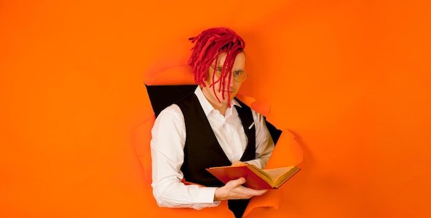 Photo homme bien habillé avec une coiffure lumineuse lisant un livre et sortant du trou de fond orange hipster impressionnant dans des lunettes avec des dreadlocks rouges lisant un vieux livre