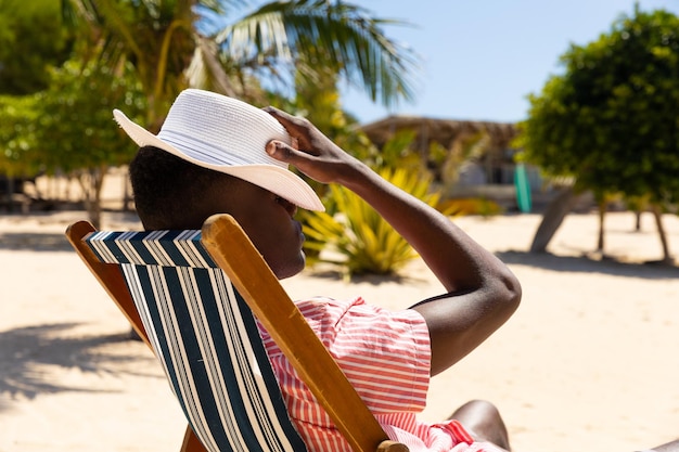 Un homme biracial détendu assis dans un fauteuil avec un chapeau sur les yeux sur une plage ensoleillée.