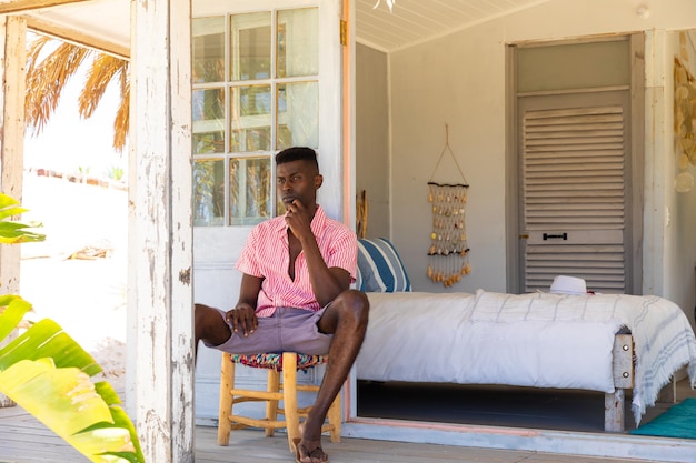 Un homme biracial réfléchi qui regarde loin assis sur le porche d'une maison d'été sur la plage, un espace de copie, un style de vie, du temps libre, de l'été et des vacances, inchangés.