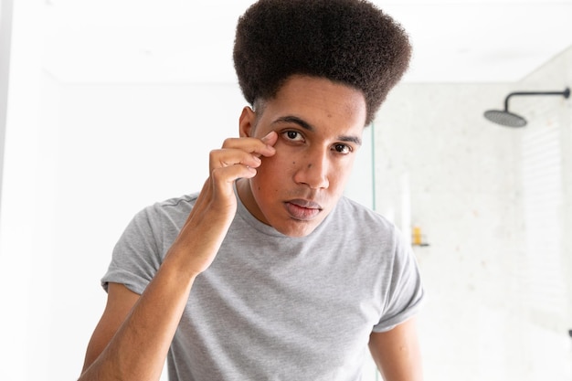 Homme biracial regardant dans un miroir inspectant les yeux dans une salle de bains ensoleillée