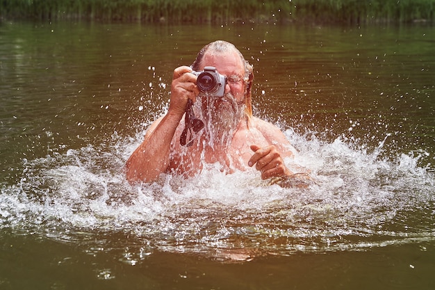 Homme blanc barbu mature nage dans la rivière et prend des photos avec un appareil photo étanche.