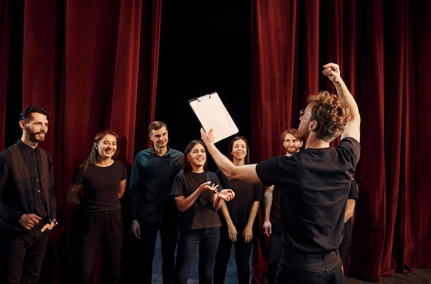 Photo un homme avec un bloc-notes pratique son rôle un groupe d'acteurs en vêtements de couleur foncée à la répétition au théâtre