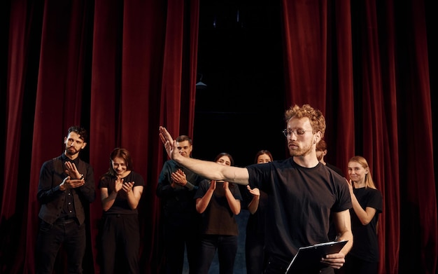 Un homme avec un bloc-notes pratique son rôle Un groupe d'acteurs en vêtements de couleur foncée à la répétition au théâtre