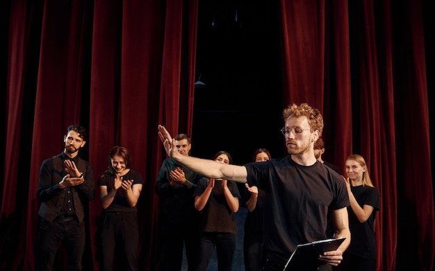 Homme avec bloc-notes pratique son rôle Groupe d'acteurs en vêtements de couleur sombre en répétition au théâtre