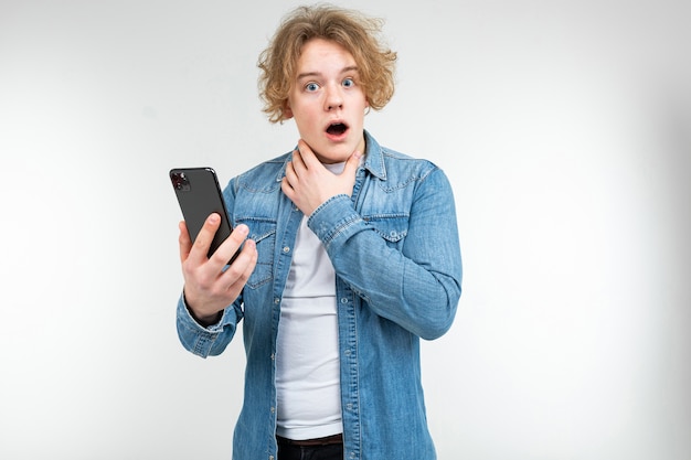 Homme blond aux cheveux blancs ondulés et surprise sur son visage avec un smartphone dans ses mains sur un fond de studio blanc.