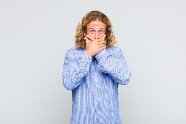 Homme blond couvrant la bouche avec les mains avec une expression choquée et surprise, gardant un secret ou disant oups