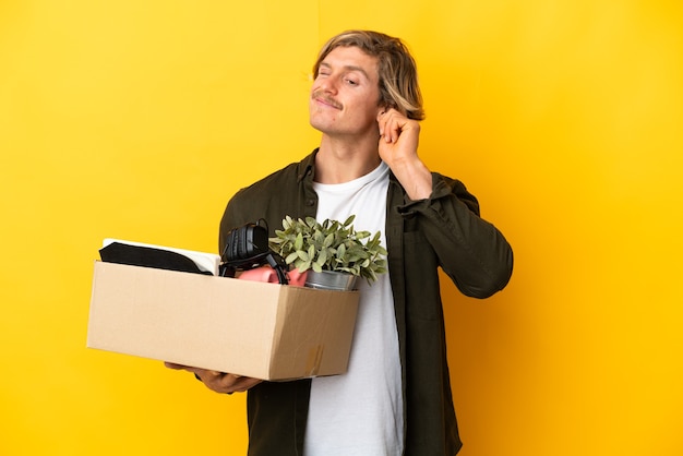 Homme blond faisant un mouvement tout en ramassant une boîte pleine de choses isolées sur fond jaune frustré et couvrant les oreilles