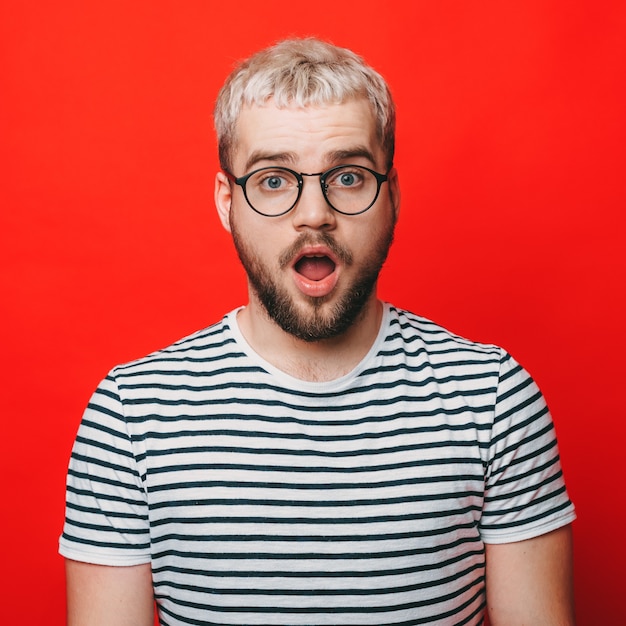 Photo homme blond gesticulant étonnement tout en portant des lunettes sur un mur de studio rouge