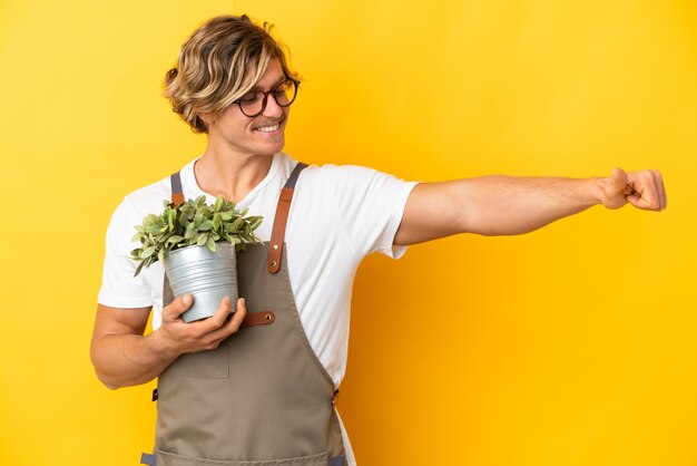 Homme blond jardinier tenant une plante isolée sur un mur jaune donnant un geste de pouce en l'air