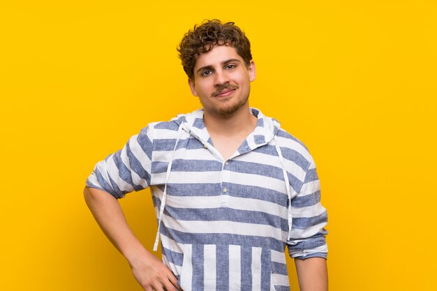 Homme blond sur un mur jaune posant avec les bras à la hanche et souriant