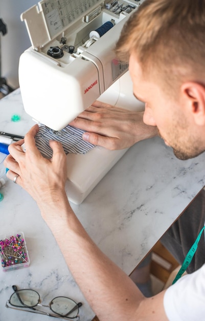 Un homme blond en t-shirt blanc coud dans un atelier à domicile Un couturier avec un ruban à mesurer sur le cou Le concept de style de vie de petite entreprise et de plaisir du travail